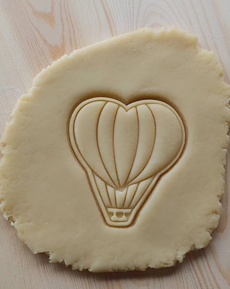 May include: A close-up of a cookie dough sheet with a hot air balloon cookie cutter pressed into it. The cookie cutter is shaped like a heart with a basket at the bottom.