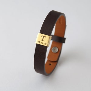 Könnte beinhalten: Ein braunes Lederarmband mit einem goldfarbenen, rechteckigen Verschluss. Der Verschluss ist mit dem Buchstaben "T" und dem Datum "05.14.20" graviert. Das Armband hat einen Druckknopfverschluss und ist für das Handgelenk konzipiert.