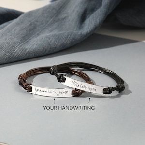 Peut inclure: Deux bracelets personnalisés avec des plaques nominatives argentées et des bracelets en cordon tressé. L'un est marron, l'autre noir. Les plaques argentées portent des textes manuscrits : "forever in my heart" et "I U Daddy Sophia".