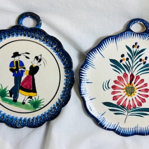 Puede incluir: Dos platos de cerámica decorativos con borde azul y blanco. Un plato presenta una escena pintada de una pareja con ropa tradicional francesa. El otro plato presenta un diseño floral pintado con una gran flor rosa.