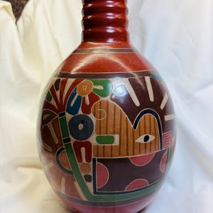 Puede incluir: Un jarrón de cerámica pintado a mano, de color terracota, con un cuerpo bulboso y un cuello rojo oscuro estriado. El jarrón presenta un diseño colorido y estilizado con formas y figuras geométricas en tonos marrón, verde, azul y rojo.