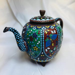 May include: A colorful, decorative teapot with a detailed floral and butterfly design. The teapot features a lid and spout, with a predominantly green, blue, and red color scheme. The teapot is supported by three small feet.