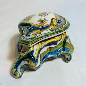 May include: A white ceramic trinket box with a lid, decorated with blue, green, and yellow floral and scroll designs. The box has a unique, three-dimensional, shell-like shape with a base that has three feet.