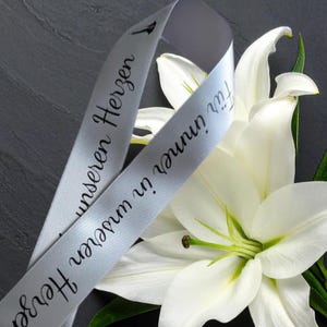 Könnte beinhalten: Nahaufnahme von weißen Lilien und einem hellgrauen Band. Das Band trägt die schwarze Aufschrift "Für immer in unseren Herzen". Die Blumen haben große, weiße Blütenblätter und grüne Stiele. Der Hintergrund ist dunkelgrau.