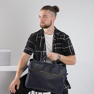 Könnte beinhalten: Eine marineblaue Lederaktentasche mit goldenem Reißverschluss und Griffen. Die Tasche hat eine rechteckige Form und einen Schulterriemen. Die Person im Bild trägt ein schwarz-weiß gemustertes Hemd und eine Uhr.