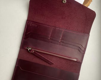 May include: Open burgundy leather wallet with multiple compartments, including card slots and a zippered pocket. The wallet is open, revealing its interior, and features a snap closure. The word "nada" is visible on a card.