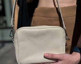 Puede incluir: Bolso bandolera rectangular color crema con superficie texturizada. El bolso tiene una correa y herrajes plateados. La palabra "soda" está grabada en la parte delantera. El bolso es sostenido por una persona.