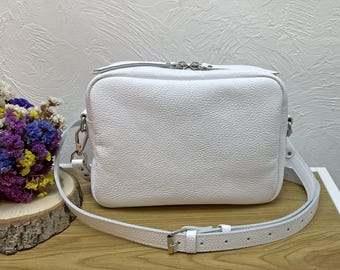Puede incluir: Un bolso bandolera blanco, rectangular, con una superficie texturizada. El bolso tiene una cremallera plateada y una correa larga y ajustable con una hebilla plateada. El bolso está sobre una superficie de madera.
