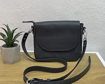 Op de afbeelding: Zwarte leren crossbodytas met een klepsluiting en verstelbare riem. De tas heeft zilverkleurige hardware en het woord "nada" in reliëf op de voorkant. De tas staat op een houten oppervlak.