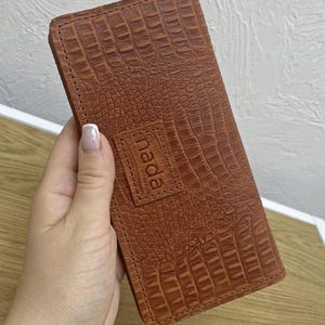 May include: A brown leather wallet with a crocodile skin texture. The wallet has a rectangular shape and a stitched border. The word "nada" is embossed on a small rectangular patch on the front.
