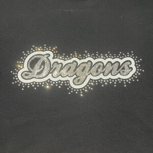 Op de afbeelding: Zwart t-shirt met het woord "Dragons" in zilveren glitterletters. Het woord is omgeven door kleine, sprankelende strass-steentjes. Het shirt heeft een ronde hals.