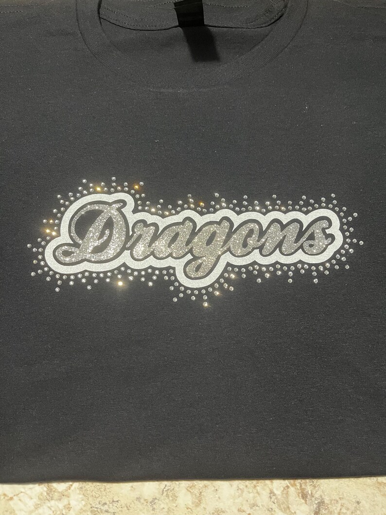 Op de afbeelding: Zwart t-shirt met het woord "Dragons" in zilveren glitterletters. Het woord is omgeven door kleine, sprankelende strass-steentjes. Het shirt heeft een ronde hals.