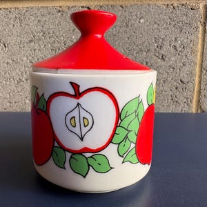 May include: A white ceramic jar with a bright red lid and apple illustrations. The jar features a large red apple with a white interior and green leaves. The lid has a rounded shape.