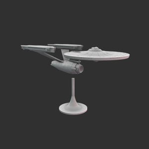 Puede incluir: Modelo gris claro de una nave espacial, con un casco principal en forma de platillo, dos góndolas de curvatura y un casco secundario. El modelo está montado sobre un soporte blanco, sobre un fondo gris oscuro.