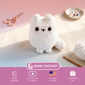 May include: A handmade white crochet cat with black eyes and pink cheeks. The cat is sitting on a light blue and white surface. The image also includes a pair of rose gold scissors, a small dish with beads, a ball of white yarn, and a crochet hook. The text "KNIRI CROCHET" is visible.