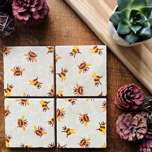 May include: Four square ceramic coasters with a cream background and a repeating pattern of yellow and brown bees. The coasters are arranged on a wooden surface, with decorative elements like a succulent and pinecones.