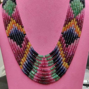 Puede incluir: Un collar de múltiples hilos con una vibrante variedad de piedras preciosas. El collar presenta un degradado de colores, incluyendo verde, rosa, amarillo, azul y negro. Las piedras están dispuestas en un patrón geométrico.