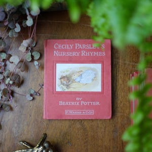 May include: A vintage copy of "Cecily Parsley's Nursery Rhymes" by Beatrix Potter. The book has a red cover with the title in white lettering and a framed illustration of a rabbit. The book is resting on a wooden surface, with greenery in the background.