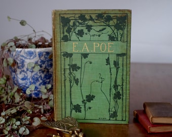 May include: A vintage book with a green cover and gold lettering that reads "E.A. POE". The cover features a botanical design. A small brass insect figurine and a small potted plant are in the foreground.
