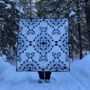 Peut inclure: Une courtepointe à motifs géométriques dans les tons de blanc, de gris et de bleu marine. La courtepointe présente un motif symétrique avec des triangles et des carrés. La courtepointe est présentée dans une forêt enneigée, avec de grands conifères en arrière-plan.