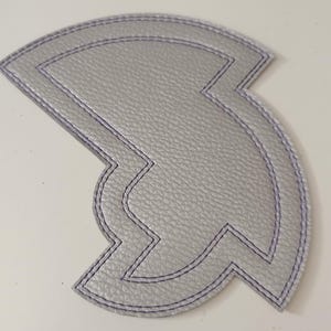 May include: A silver-coloured, leather-like patch with a unique, geometric design. The patch has a stitched outline in a contrasting purple colour. The shape is a stylized, asymmetrical design, possibly for a decorative application.