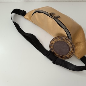 Puede incluir: Una riñonera de cuero dorada con una correa ajustable negra y un parche de cuero marrón con el texto "A l'endroit Couture" en puntadas doradas.