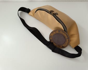 May include: A gold leather fanny pack with a black adjustable strap and a brown leather patch with the text "A l'endroit Couture" in gold stitching.