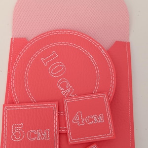 Può includere: Una custodia color corallo con patta rosa chiaro e un bottone a pressione bianco. Contiene un pezzo circolare etichettato 10 cm, un pezzo quadrato da 5 cm, un pezzo quadrato da 4 cm e un pezzo quadrato più piccolo etichettato 3 cm.