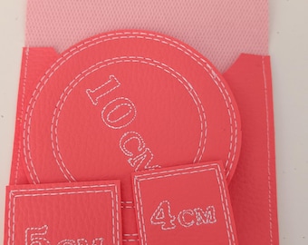 Op de afbeelding: Een koraalkleurig envelop-etui met een lichtroze flap en een witte drukknoopsluiting. Het bevat een cirkelvormig onderdeel met 10 cm, een vierkant onderdeel met 5 cm, een vierkant onderdeel met 4 cm en een klein onderdeel met 3 cm.