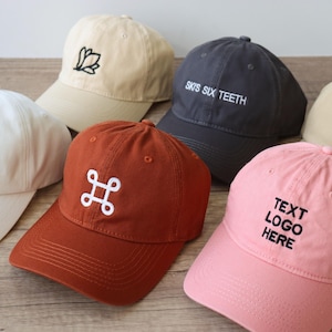 Op de afbeelding: Een verzameling baseballpetten in diverse kleuren, waaronder wit, beige, roest, marineblauw en roze. Elke pet heeft geborduurde tekst of een logo in contrasterende kleuren. De petten liggen op een houten oppervlak.