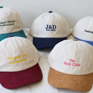 May include: Five corduroy baseball caps with cream-colored crowns and colored brims. Each cap has embroidered text: "Hobart Owens Invitational", "J&D Est.2025", "Redding's Dad", "bad girls are modernists", and "The Bach Club".