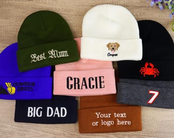May include: A collection of beanies in various colors, including blue, green, pink, black, and brown. Some beanies feature embroidered text like "Best Mum," "Gracie," and "BIG DAD." Others have graphics such as a crab, a dog, and the number 7.