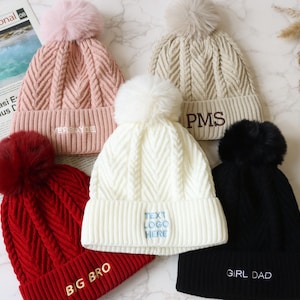 May include: Five knitted bobble hats in a variety of colours, including pink, cream, red, white, and black. Each hat features a faux fur pom-pom and embroidered text: "VERSAYOE", "PMS", "BIG BRO", "TEXT LOGO HERE", and "GIRL DAD".