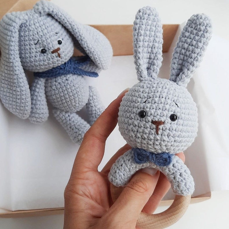 May include: Two crocheted stuffed animals, a bunny and a rabbit, both gray with blue accents. The bunny is holding a wooden teething ring.