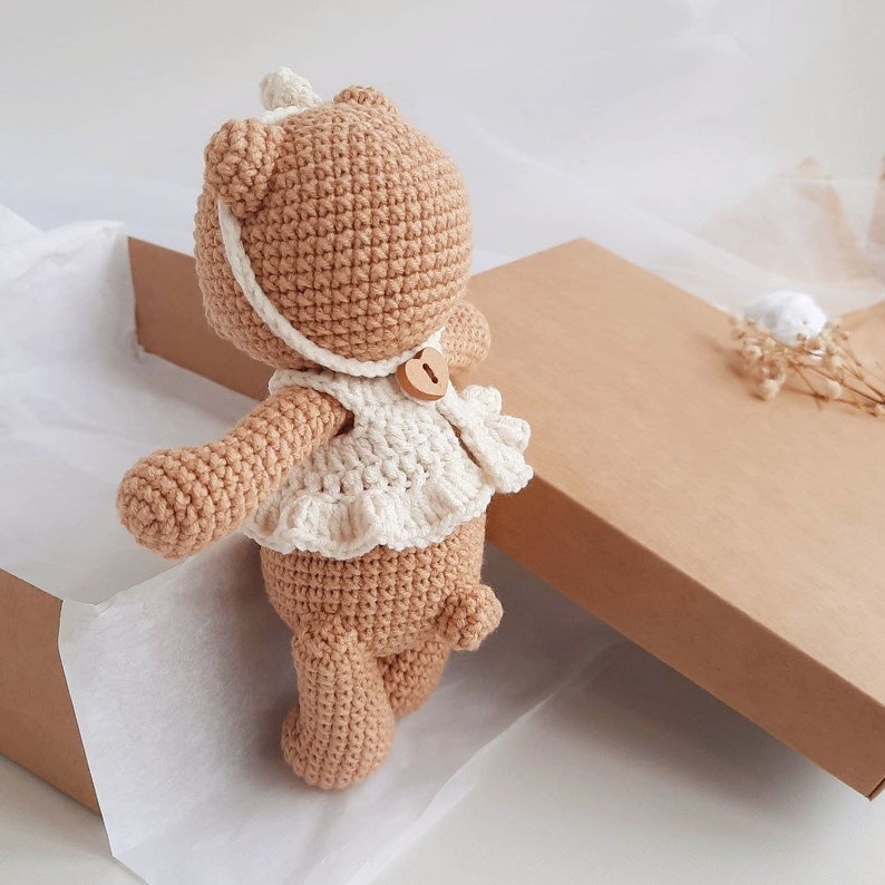Puede incluir: Un oso de peluche de crochet marr&oacute;n que lleva un vestido blanco de crochet con un bot&oacute;n de madera. El oso est&aacute; de pie en una caja marr&oacute;n con papel de seda blanco.