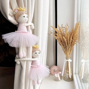 Op de afbeelding: Twee gehaakte ballerinapoppen met roze tutu's en gouden kronen worden gebruikt als gordijnbandjes op witte gordijnen. De poppen houden de gordijnen vast met hun armen.