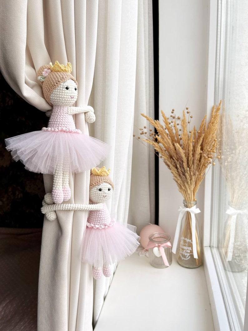 Op de afbeelding: Twee gehaakte ballerinapoppen met roze tutu's en gouden kronen worden gebruikt als gordijnbandjes op witte gordijnen. De poppen houden de gordijnen vast met hun armen.