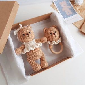 Puede incluir: Dos osos de peluche de crochet de color marr&oacute;n, uno con un vestido blanco y el otro con un babero blanco, est&aacute;n en una caja marr&oacute;n con papel de seda blanco. El oso m&aacute;s peque&ntilde;o sostiene un sonajero de madera.