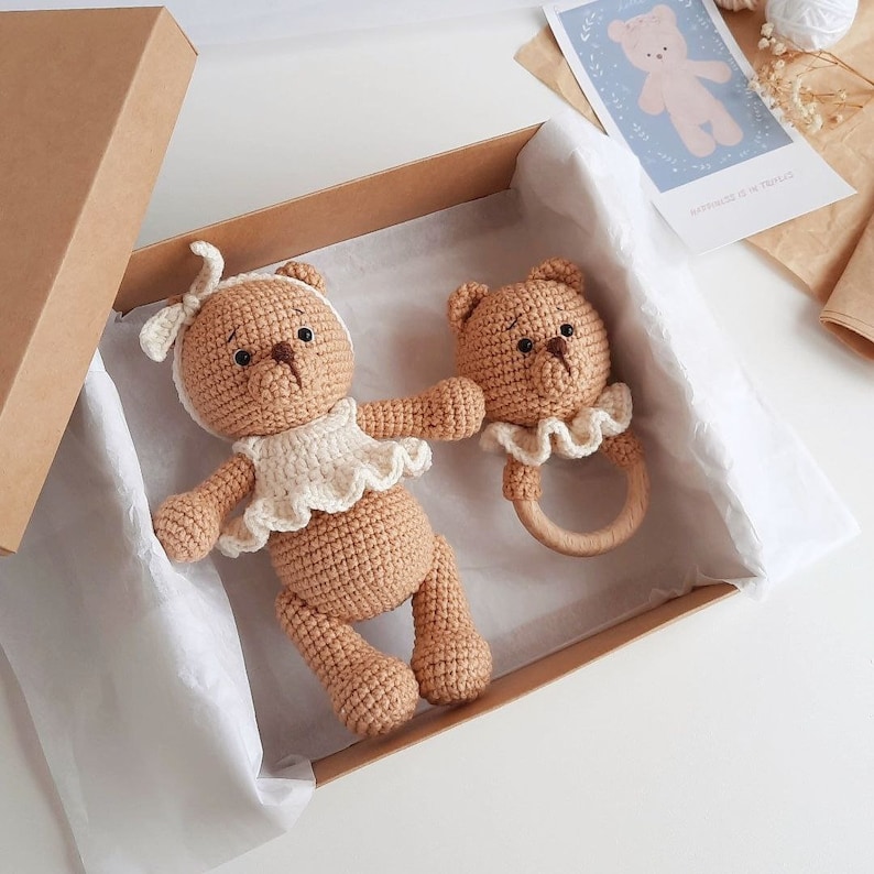 Puede incluir: Dos osos de peluche de crochet de color marr&oacute;n, uno con un vestido blanco y el otro con un babero blanco, est&aacute;n en una caja marr&oacute;n con papel de seda blanco. El oso m&aacute;s peque&ntilde;o sostiene un sonajero de madera.