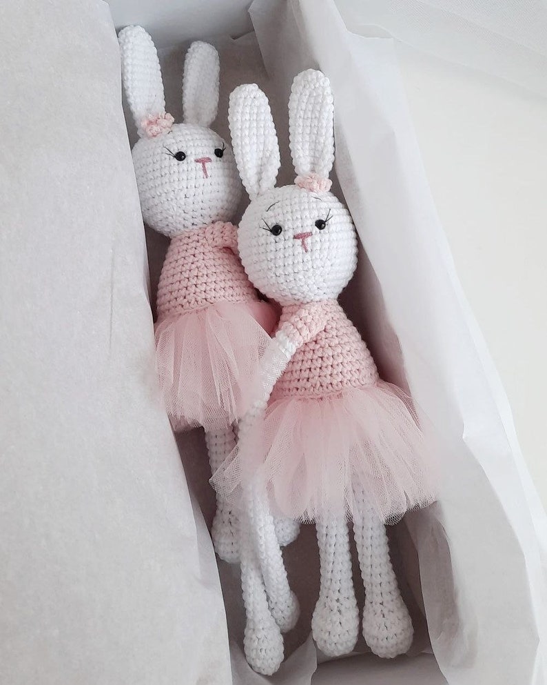 May include: Two white crocheted bunny dolls wearing pink tutus. The bunnies have black eyes and pink noses. They are sitting in a white gift box.