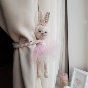 Puede incluir: Un conejito de bailarina rosa tejido a crochet con una falda de tul se utiliza como sujeta cortinas. El conejito se agarra a una cortina beige con un sujeta cortinas de cuerda.