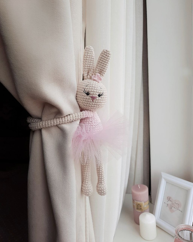 Puede incluir: Un conejito de bailarina rosa tejido a crochet con una falda de tul se utiliza como sujeta cortinas. El conejito se agarra a una cortina beige con un sujeta cortinas de cuerda.