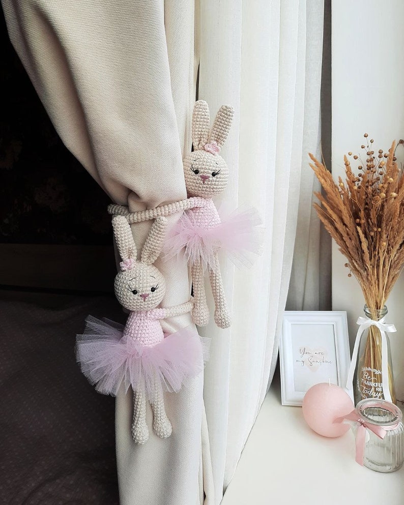 May include: Two crocheted bunny rabbit curtain tiebacks with pink tutus. The bunnies are holding onto a white curtain. The bunnies are facing the camera.