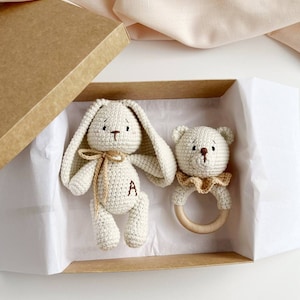 May include: A crochet bunny and bear rattle set in a gift box. The bunny has long ears, a bow, and the letter "A" on its body. The bear rattle has a wooden ring and a ruffled collar. The set is in a brown box with white tissue paper.