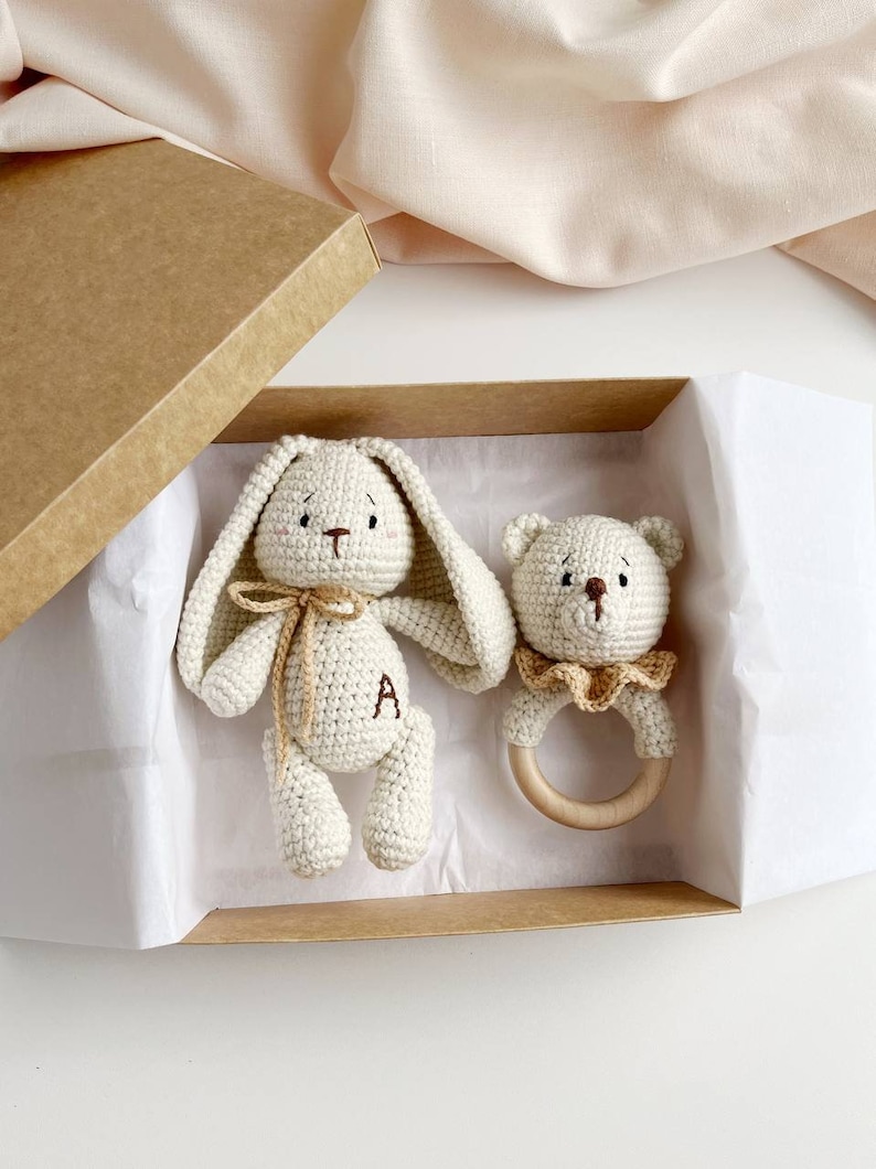 May include: A crochet bunny and bear rattle set in a gift box. The bunny has long ears, a bow, and the letter "A" on its body. The bear rattle has a wooden ring and a ruffled collar. The set is in a brown box with white tissue paper.
