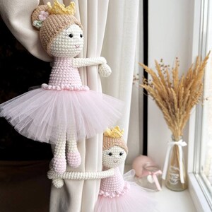 Op de afbeelding: Twee gehaakte ballerinapoppen met roze tutu's en gouden kronen worden gebruikt als gordijnbandjes. De poppen houden een wit gordijn vast.