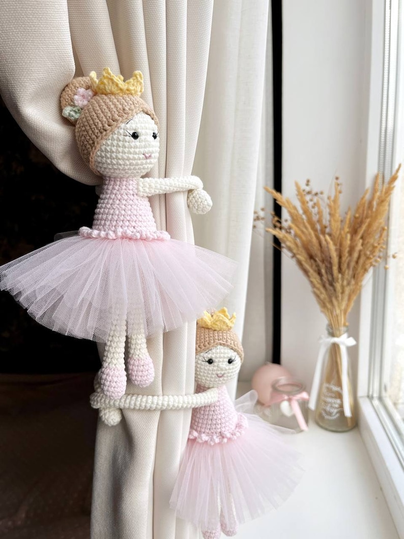 Op de afbeelding: Twee gehaakte ballerinapoppen met roze tutu's en gouden kronen worden gebruikt als gordijnbandjes. De poppen houden een wit gordijn vast.