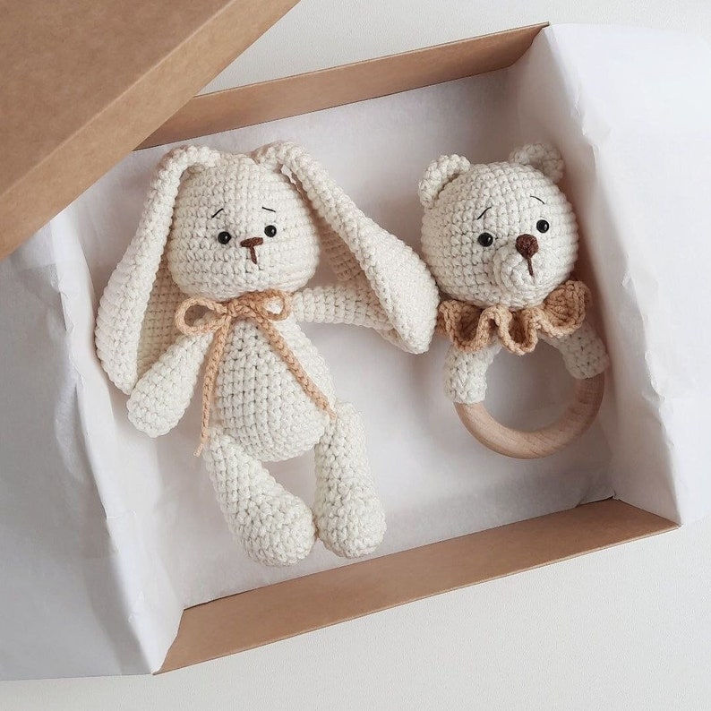 May include: A white crocheted bunny and a white crocheted bear rattle in a brown box with white tissue paper. The bear rattle has a wooden ring.