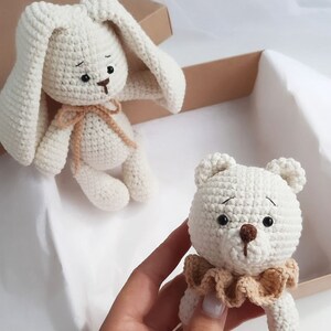 May include: Two crocheted stuffed animals, a white bunny and a white bear, both with brown eyes and noses. The bunny has long ears and a brown ribbon around its neck. The bear has a brown ruffle around its neck.