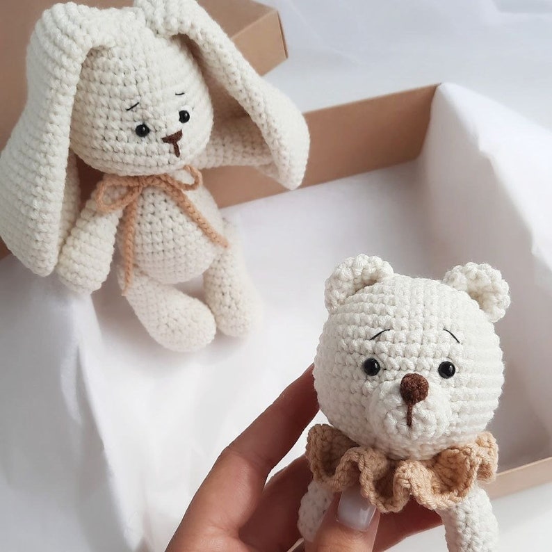 May include: Two crocheted stuffed animals, a white bunny and a white bear, both with brown eyes and noses. The bunny has long ears and a brown ribbon around its neck. The bear has a brown ruffle around its neck.